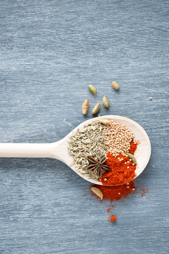 Top view of spices including star anise and chili powder in a wooden spoon on a textured blue surface.