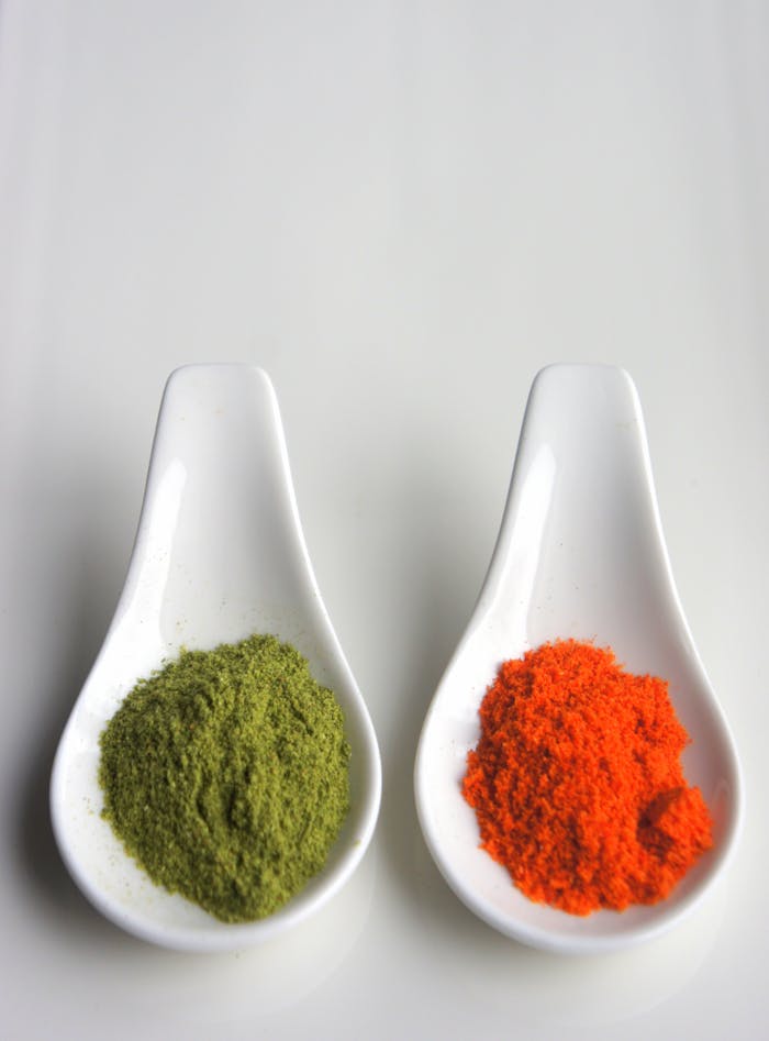 Two ceramic spoons with vibrant green and red spice powders on a neutral background.